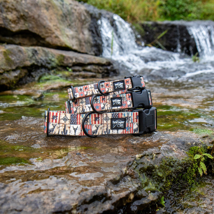 Three rugged dog collars with a distressed American flag print are stacked on wet rocks in a shallow stream with a small waterfall in the background.