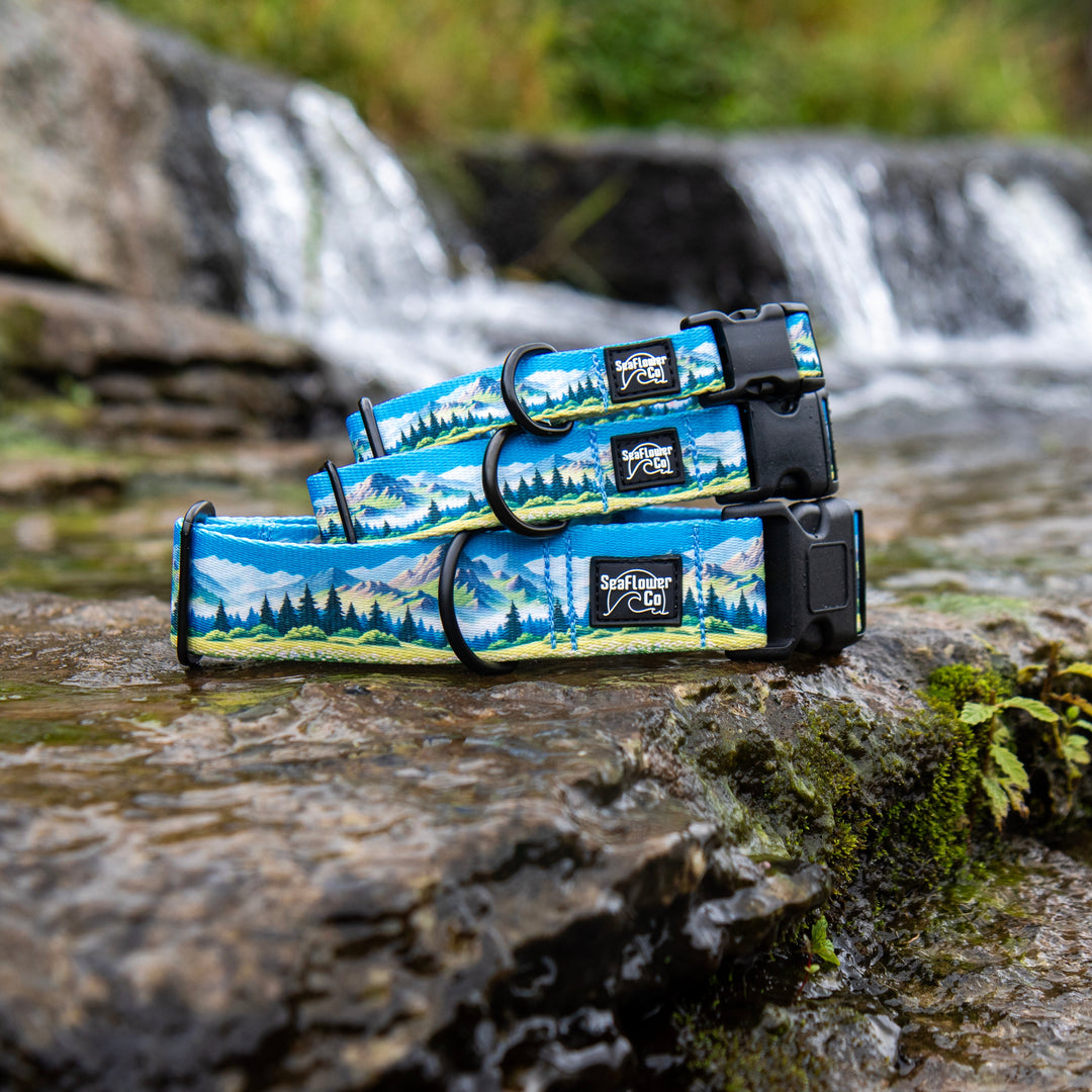Three blue outdoor dog collars with a mountain landscape print are arranged on a wet rock in a stream with a waterfall blurred in the background.