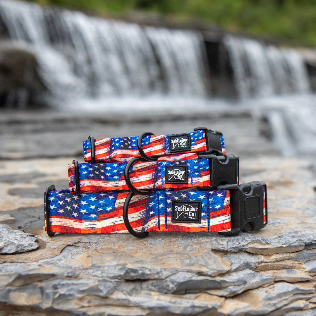 Three dog collars with an American flag print are stacked on a flat rock with a blurred waterfall in the background.