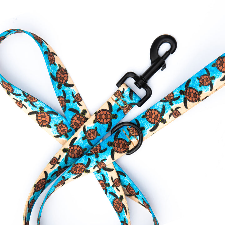 A close-up of a dog leash with a repeating sea turtle pattern on a light beige background, featuring bright blue water motifs and black hardware clasp.